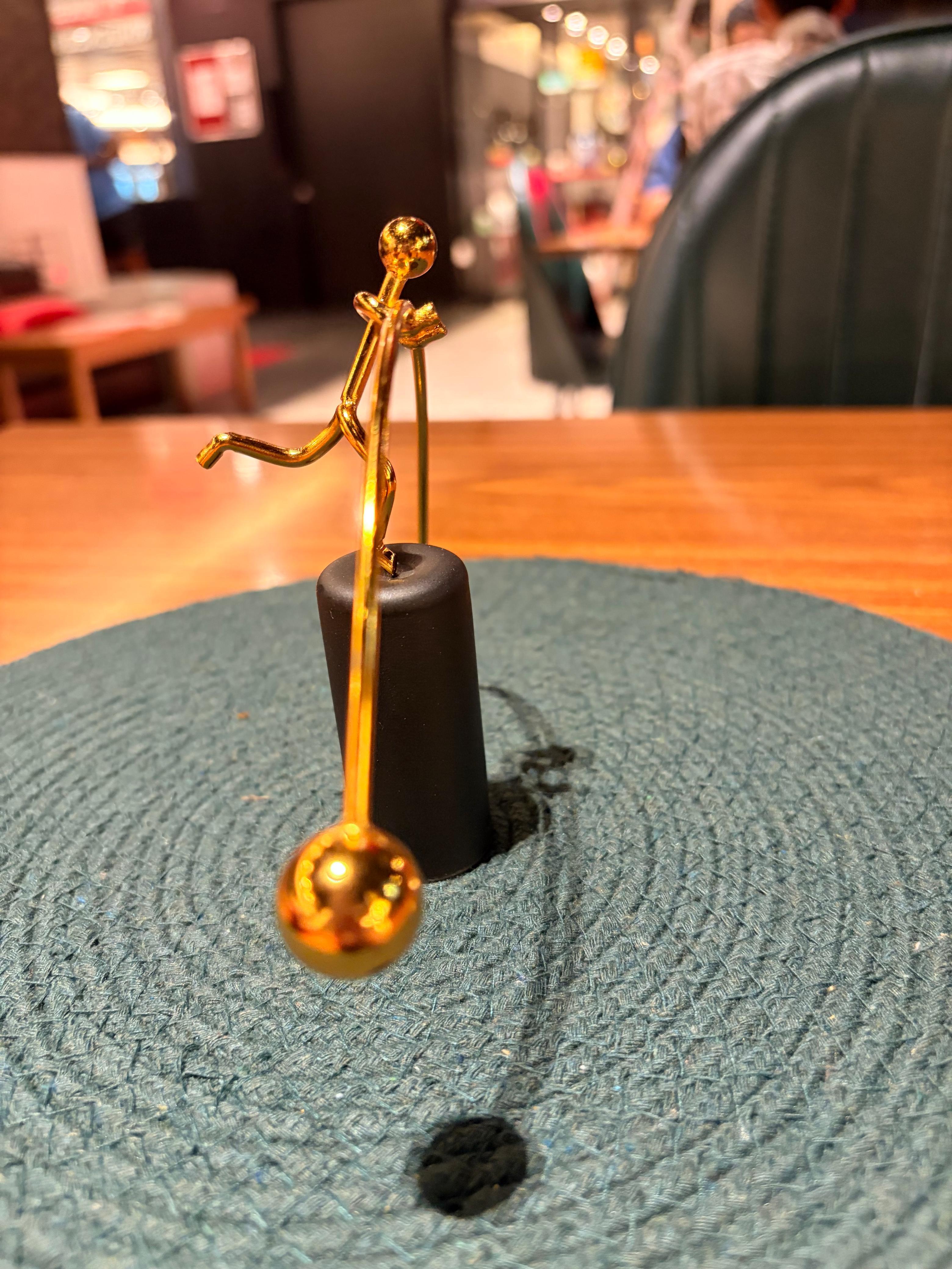 A balancing figurine — a figure balanced on one leg holding a counterweight rod, representing BALANCE+'s philosophy